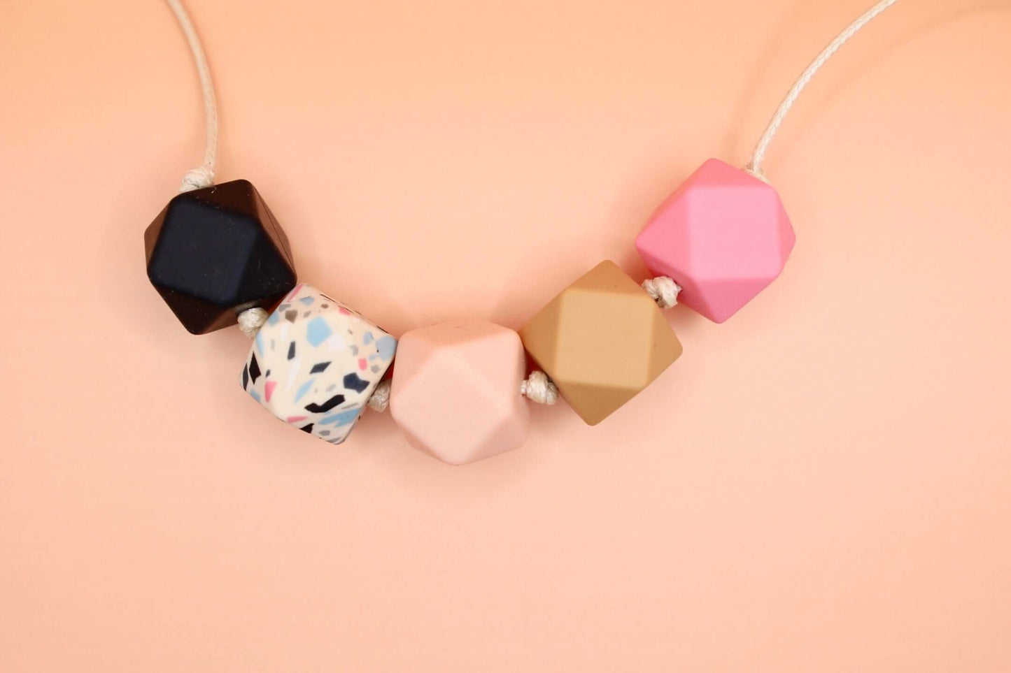 Hexagon silicone necklace with black, white speckled, pink, caramel, and pink beads on a cream cord against a peach background