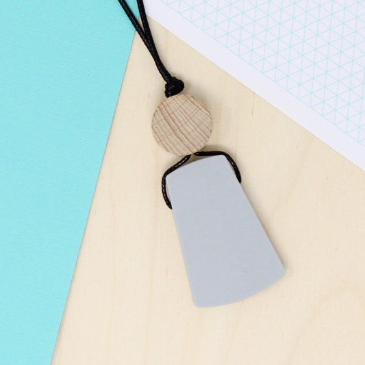 Light grey trapezoid shaped silicone pendant necklace laying flat and photographed from above on a contrasting turquoise and natural wood background.