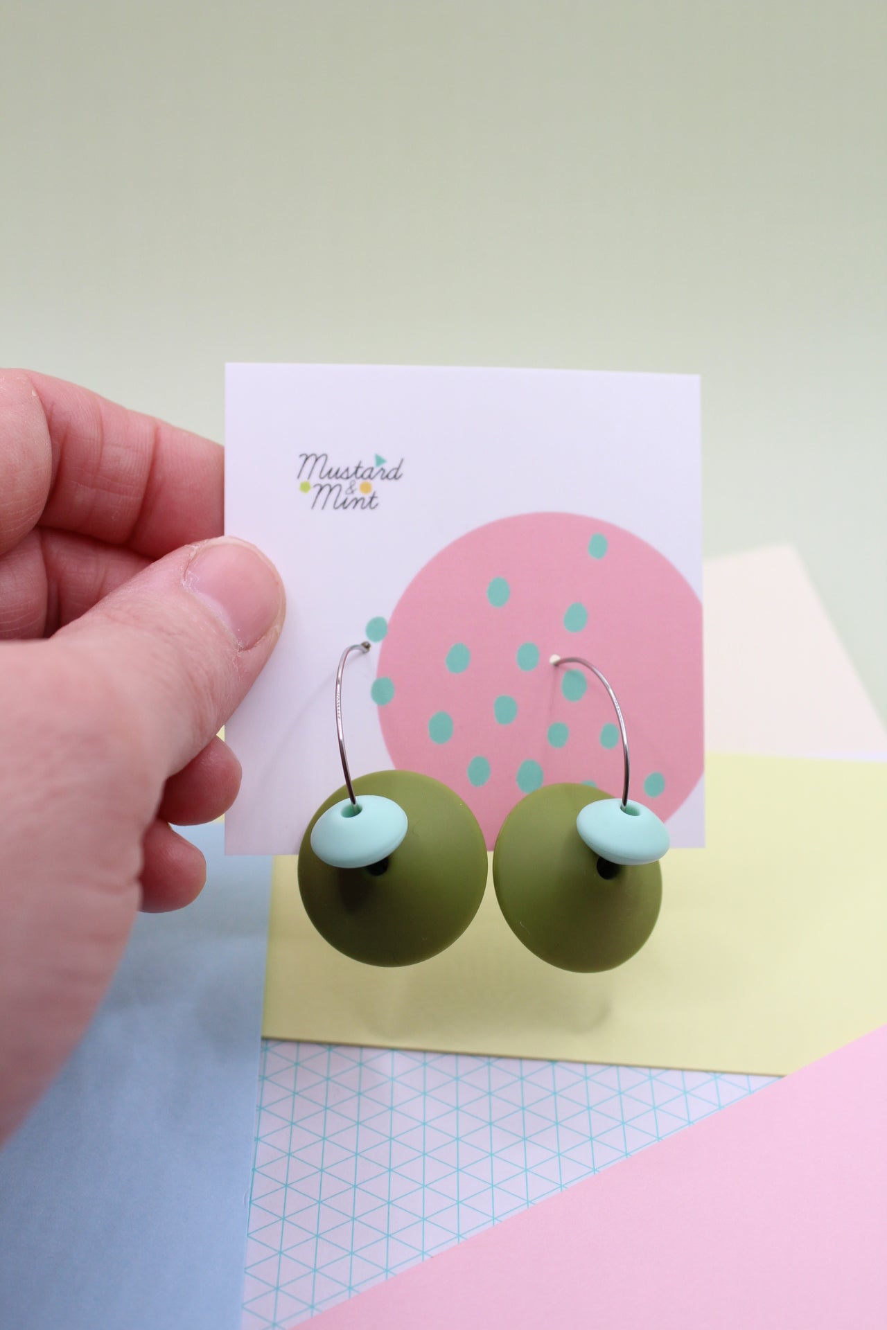 Army Green silicone dangle earrings being held up in models fingers on their Mustard & Mint branded backing board.