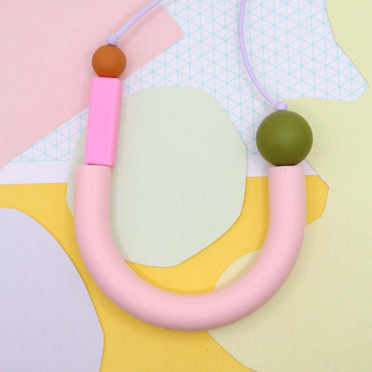 Pink U shaped silicone necklace photographed laying flat from above against mixed pastel paper background.