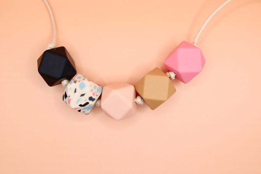 Hexagon silicone necklace with black, white speckled, pink, caramel, and pink beads on a cream cord against a peach background